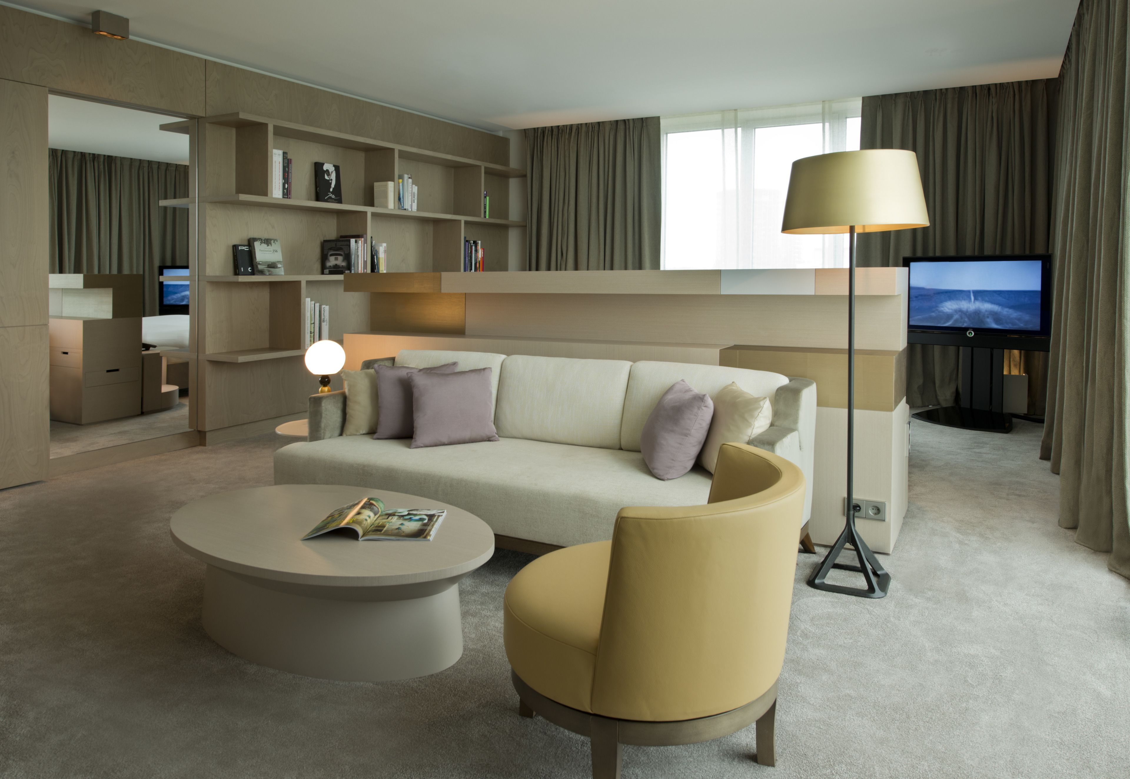 A modern hotel room with a sofa, armchair, coffee table, TV, bookshelf, and a large window with curtains.