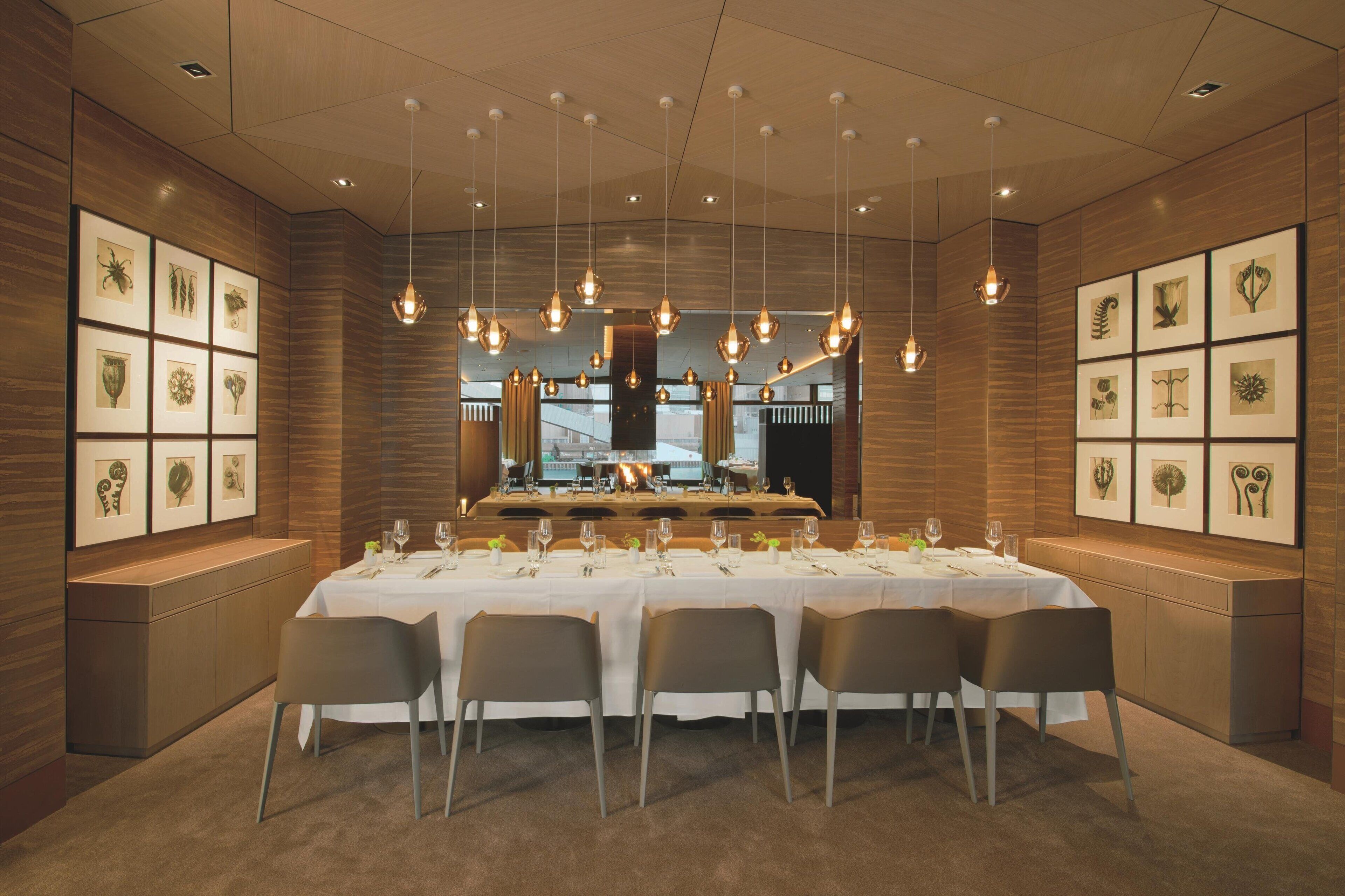 A dining area with a long table set for six, surrounded by chairs, with hanging pendant lights and framed botanical artwork on the walls.