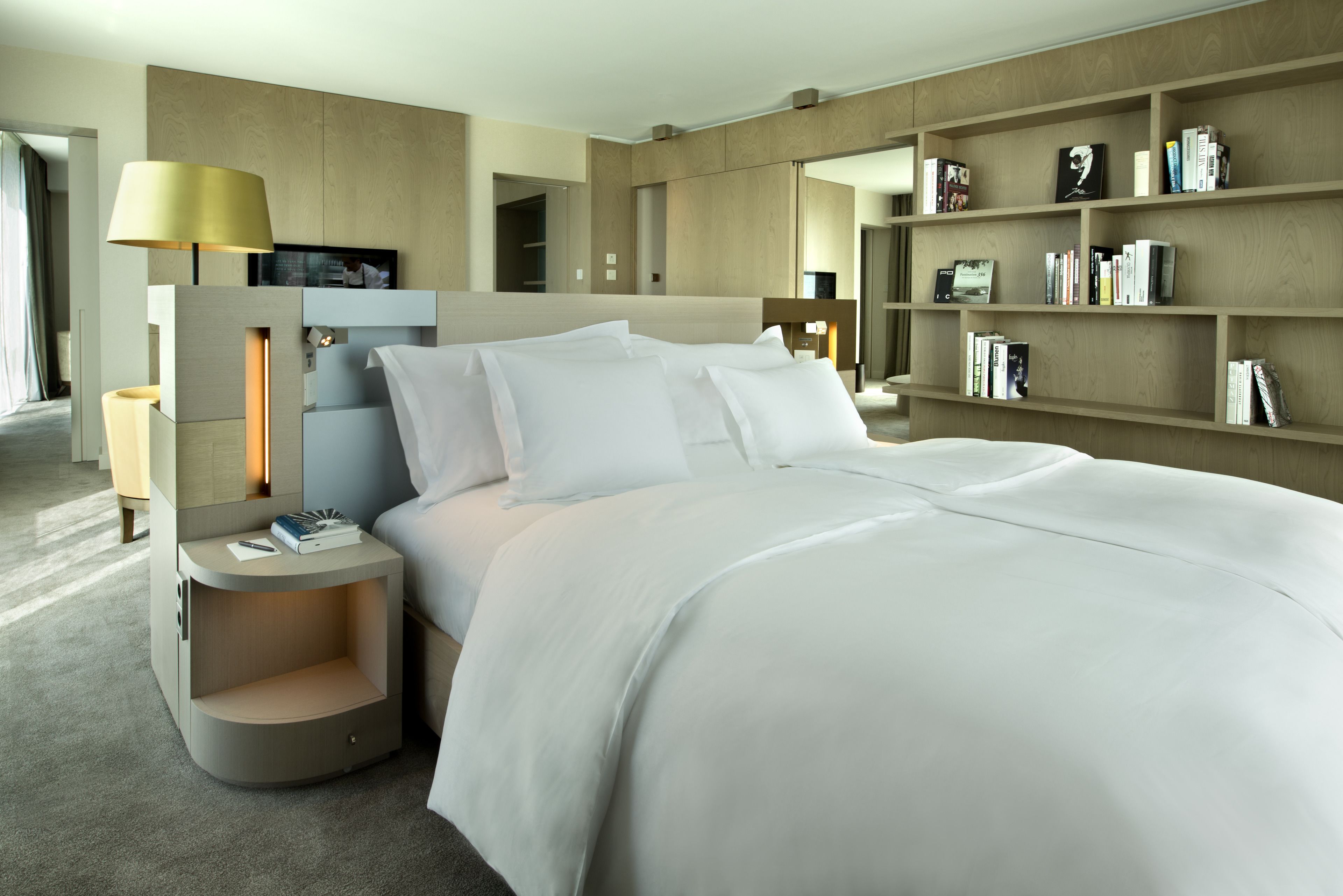 A modern hotel room with a large bed, a bedside table with a lamp, a bookshelf, and a TV on a stand.