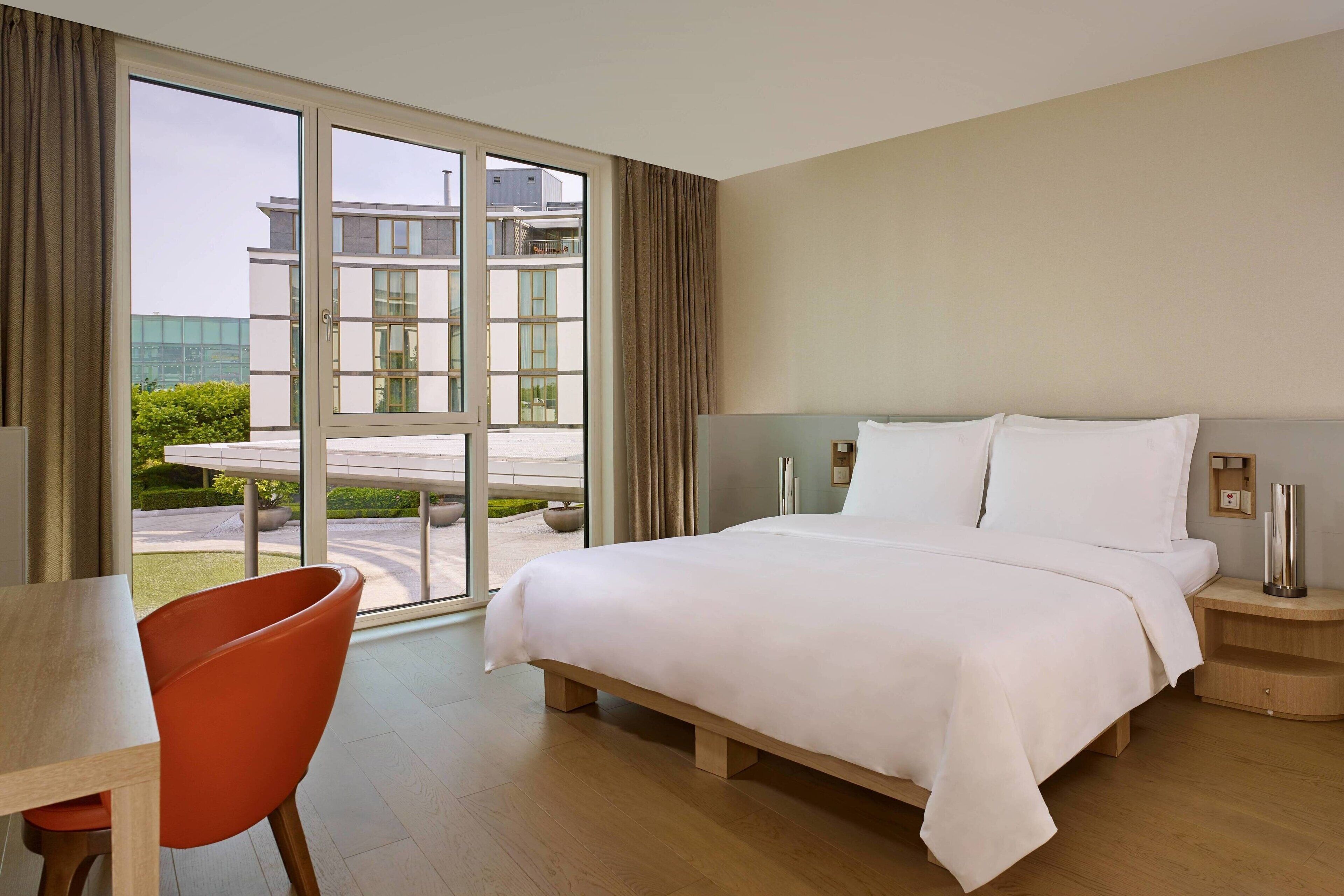 Ein Hotelzimmer mit einem großen Bett, einem Holztisch, einem roten Sessel und Blick auf ein Gebäude und Grünanlagen durch eine Glastür.