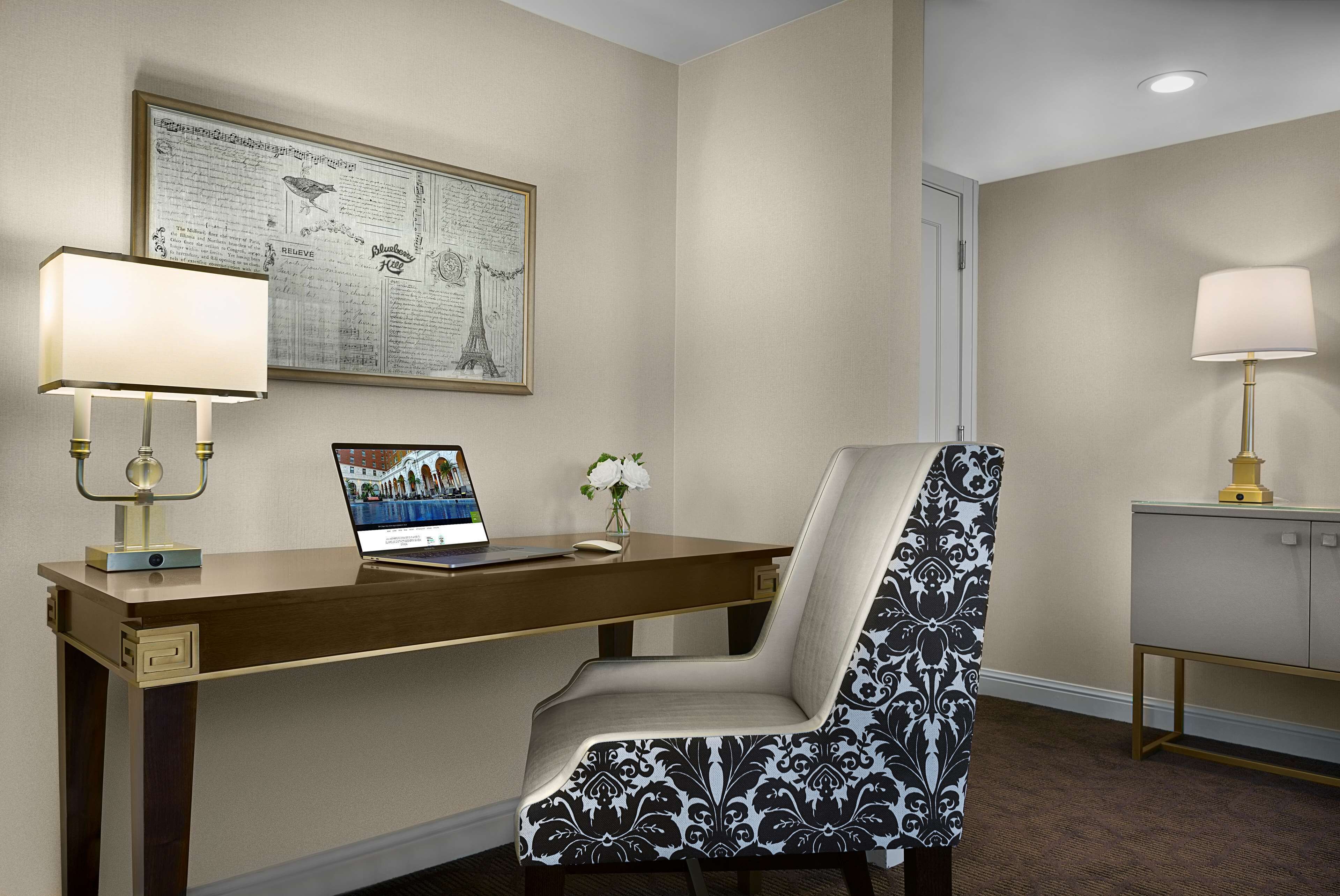 A hotel room with a desk, a laptop, a chair, and a lamp.