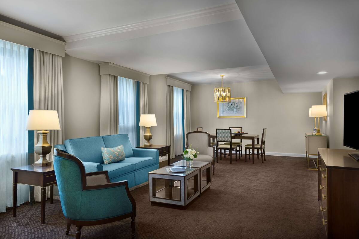 Une chambre d’hôtel avec un canapé bleu, une table basse, un téléviseur, une table à manger avec des chaises et un lustre.
