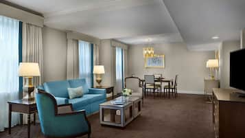 A hotel room with a blue sofa, a coffee table, a TV, a dining table with chairs, and a chandelier.