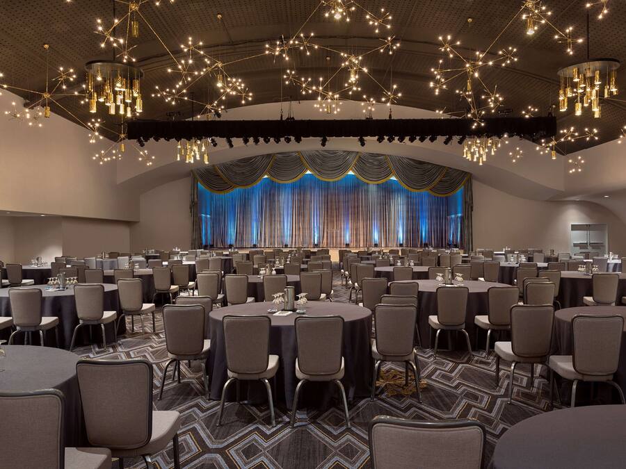 A large conference room with round tables and chairs, a stage with curtains, and a ceiling with hanging lights.