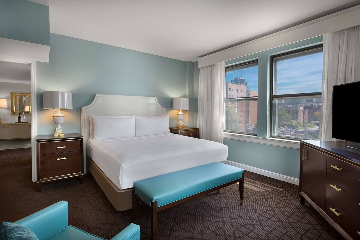 Une chambre d’hôtel avec un grand lit, deux tables de chevet avec des lampes, un banc, une télévision et une fenêtre donnant sur la ville.