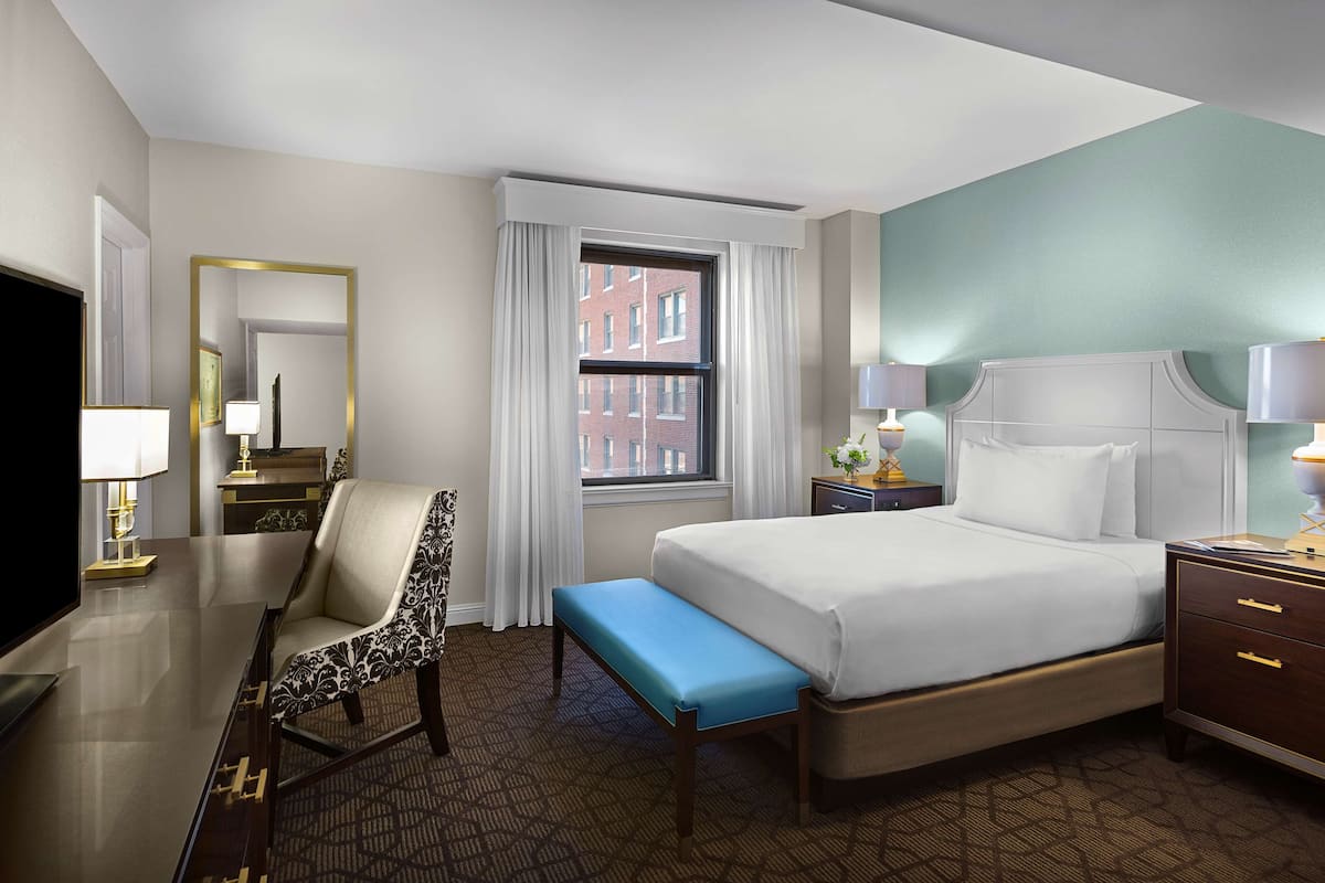 Une chambre d’hôtel comprenant un lit, un bureau avec une chaise, un banc, une table de chevet et un téléviseur.
