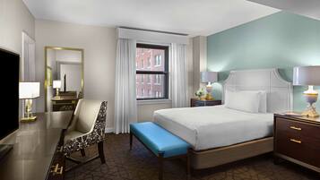 A hotel room with a bed, a desk with a chair, a bench, a nightstand, and a TV.