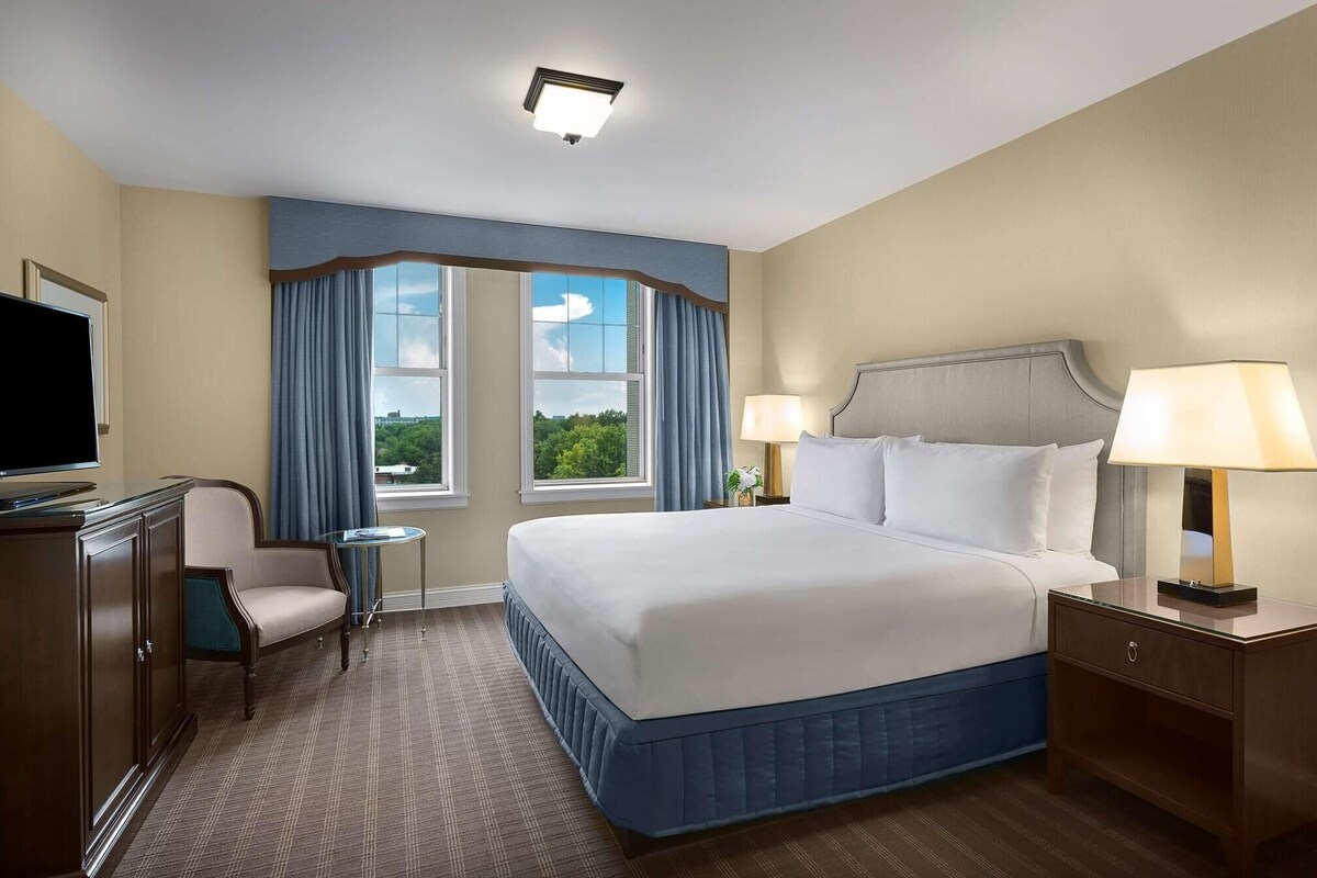 A hotel room with a large bed, a desk with a TV, a chair, two windows with curtains, and a nightstand with a lamp.
