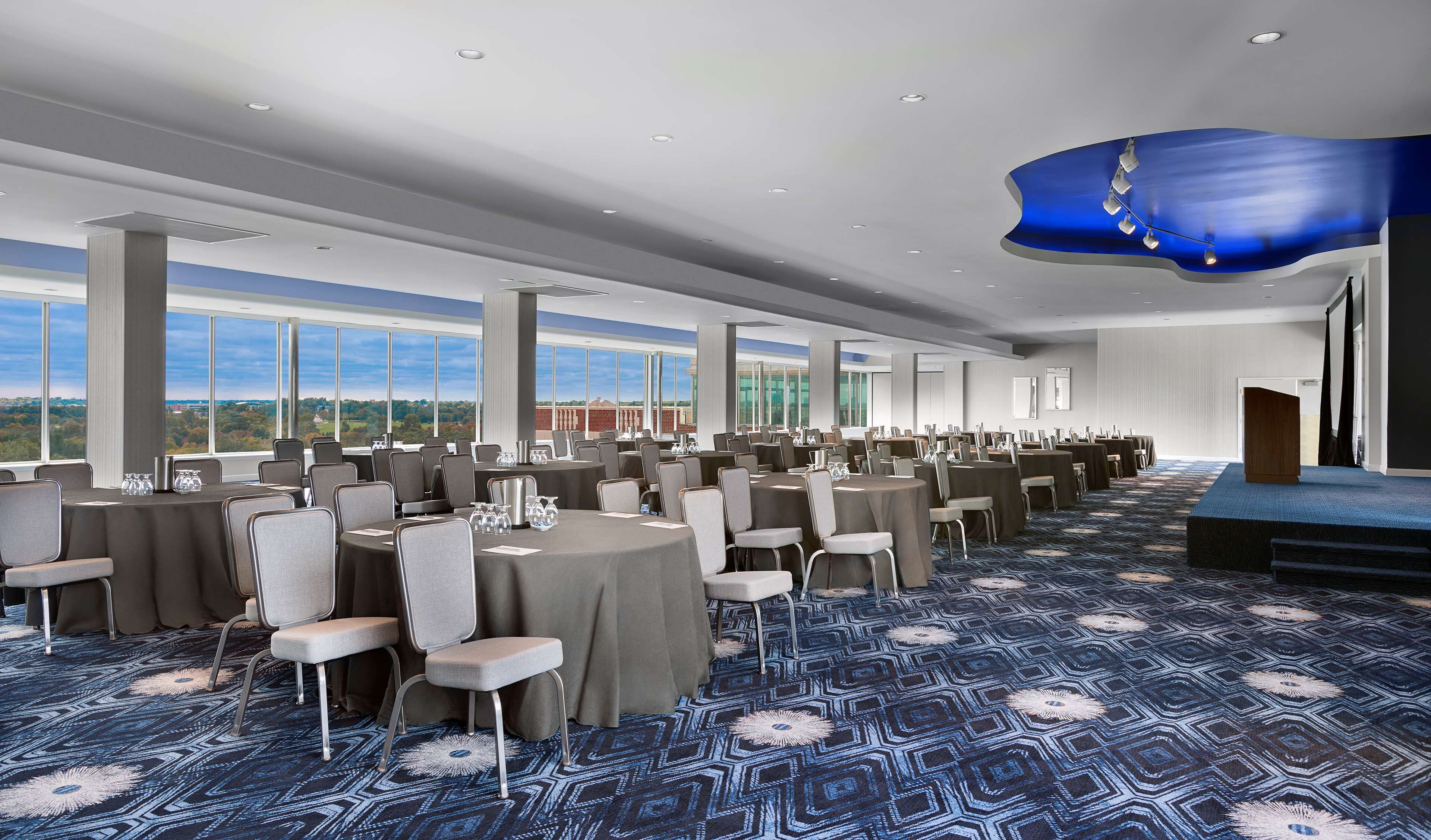 A conference room with round tables and chairs, large windows offering a view of the landscape, and a stage with a blue backdrop.