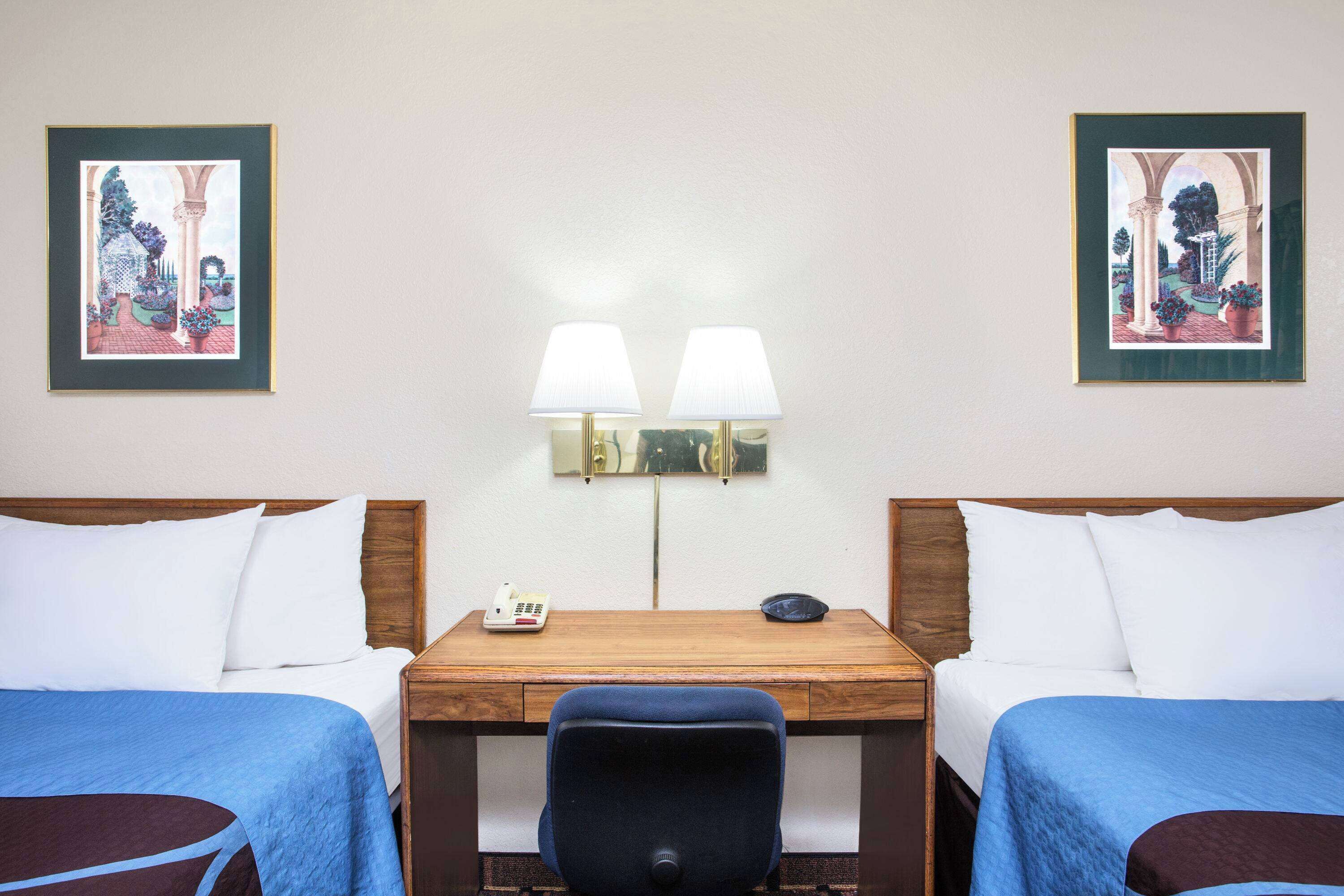 A hotel room with two beds, a desk, two lamps, and two framed pictures on the wall.