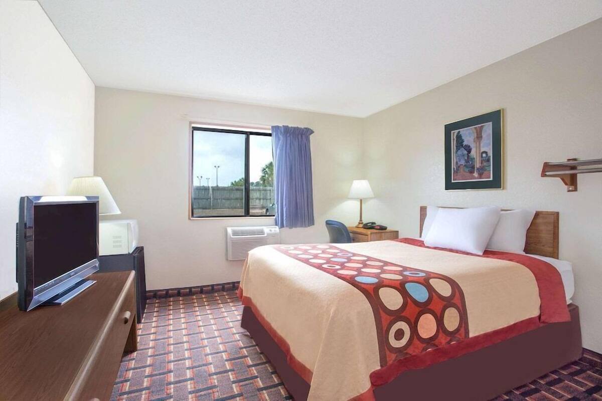Una habitación de hotel con cama, escritorio, silla, televisor y ventana con cortinas.