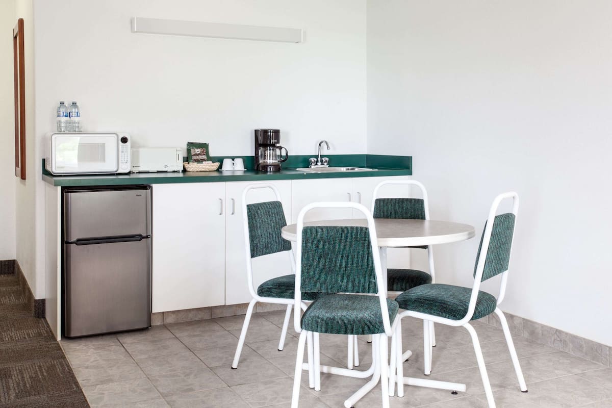 Une cuisine compacte équipée d’un réfrigérateur, d’un four à micro-ondes, d’une machine à café et d’un évier. Quatre chaises à revêtement vert sont disposées autour d’une petite table ronde.