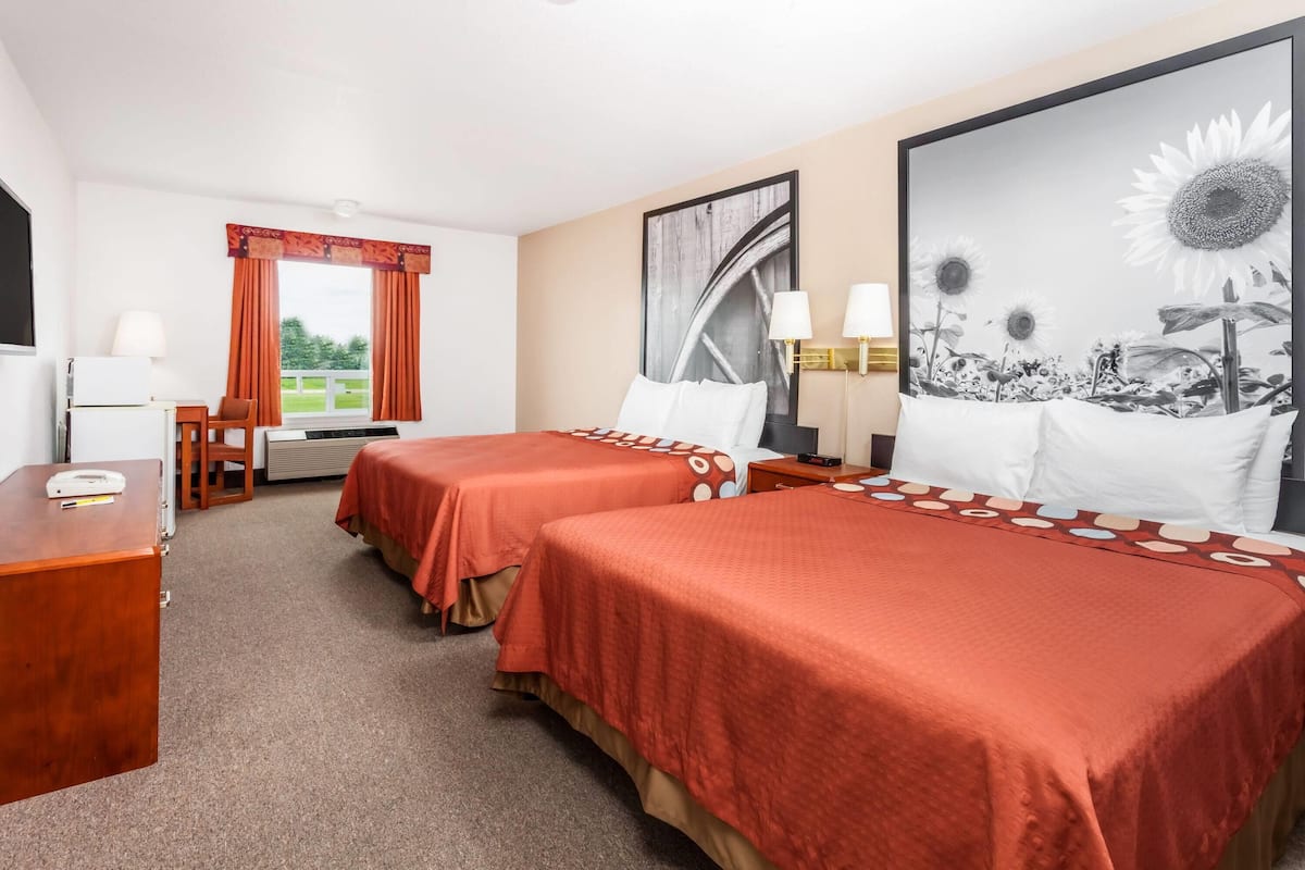 Une chambre d’hôtel avec deux lits, un bureau, une chaise, une fenêtre avec des rideaux, un tableau représentant des tournesols et une lampe.