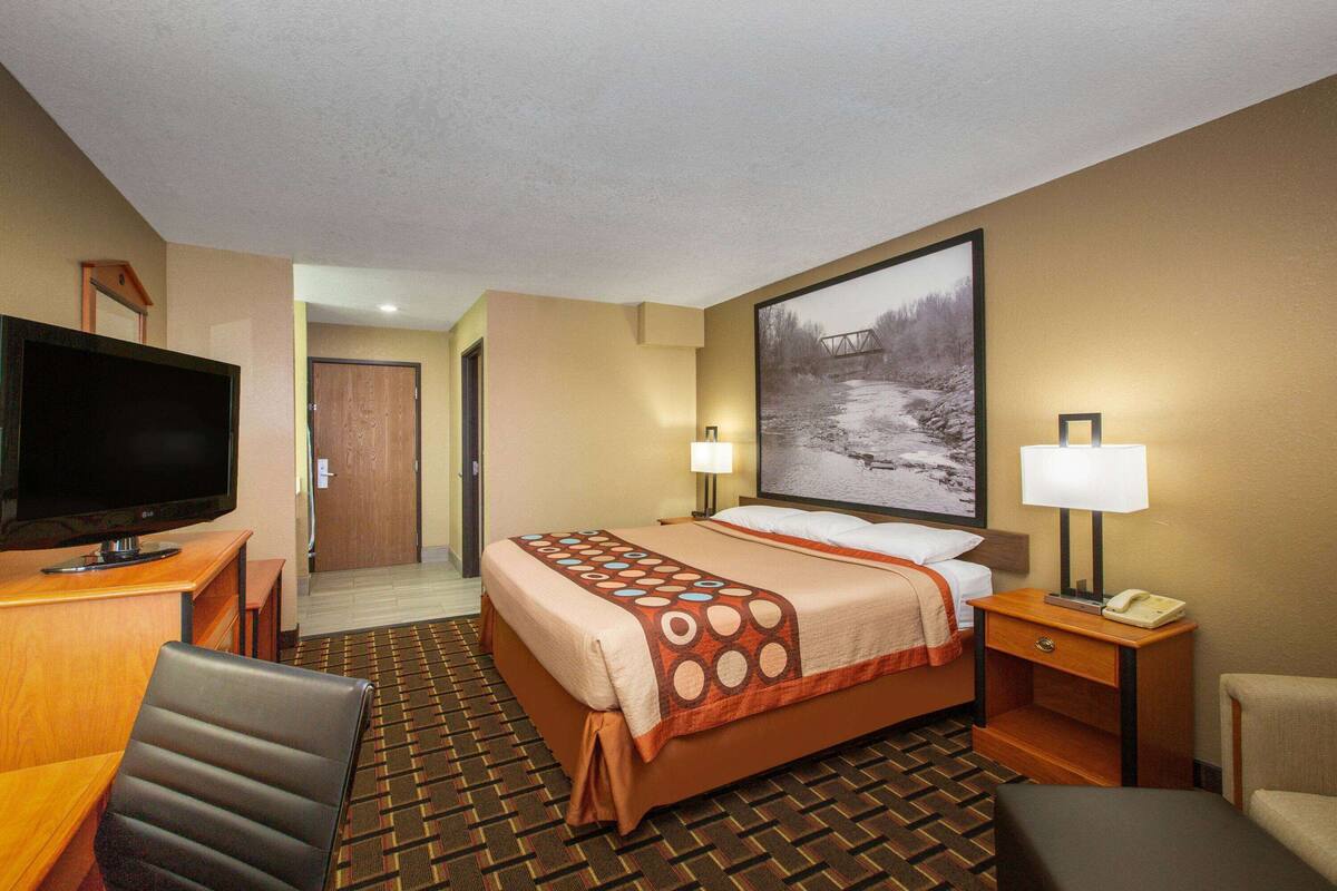 A hotel room with a bed, a desk with a TV, a chair, a lamp, and a painting on the wall.