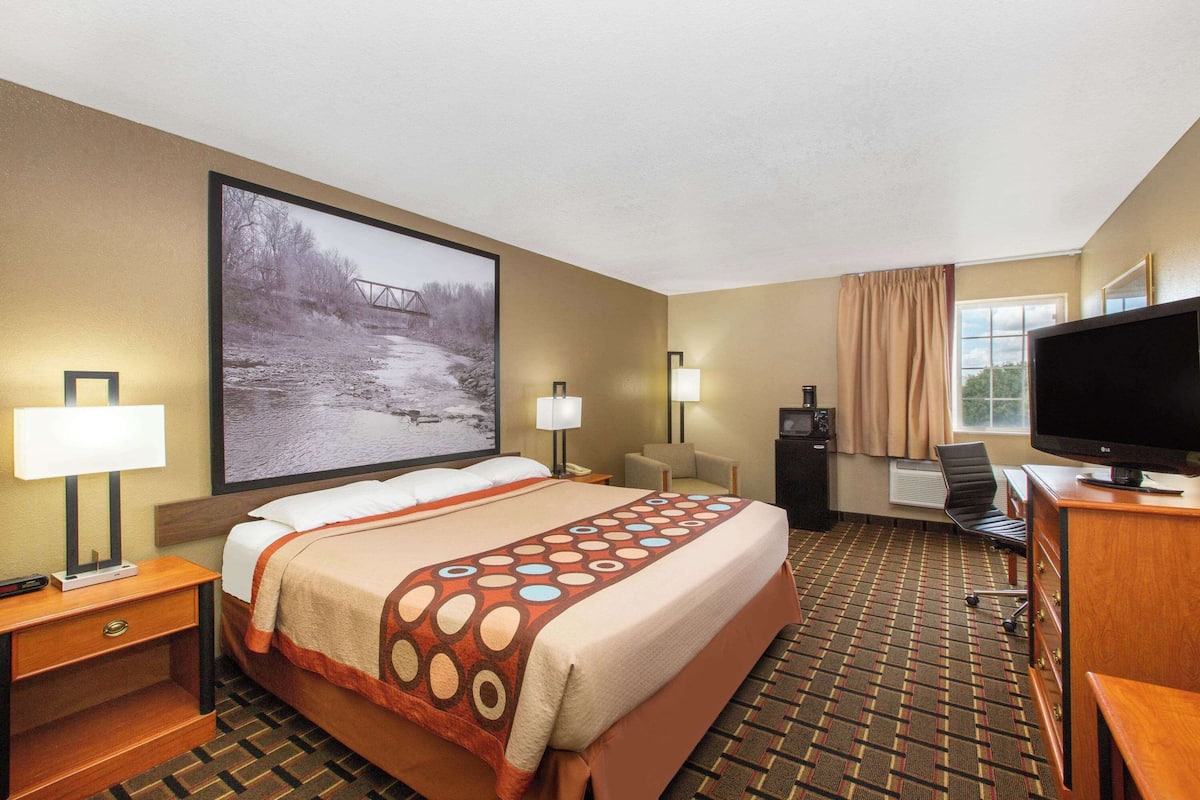 A hotel room with a large bed, two bedside tables with lamps, a flat-screen TV, a desk with a chair, and a large window with curtains.