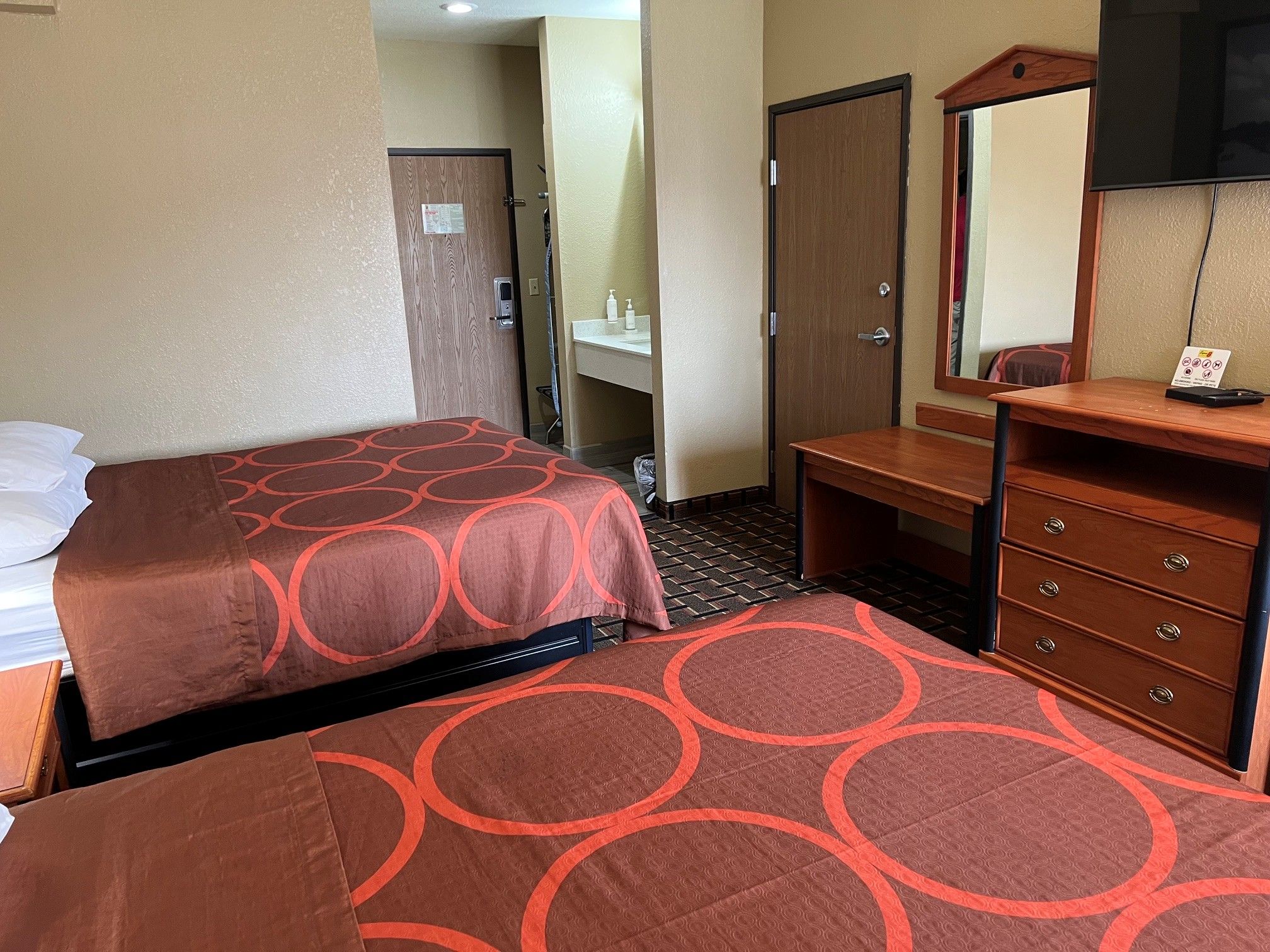 Habitación de hotel con dos camas, un aparador de madera, un espejo y un baño con lavamanos y toallero.