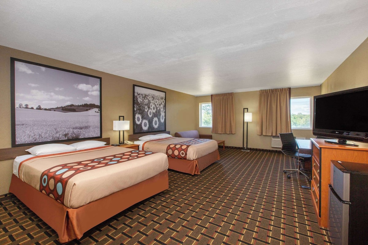 A hotel room with two beds, a desk with a TV, and a large window.