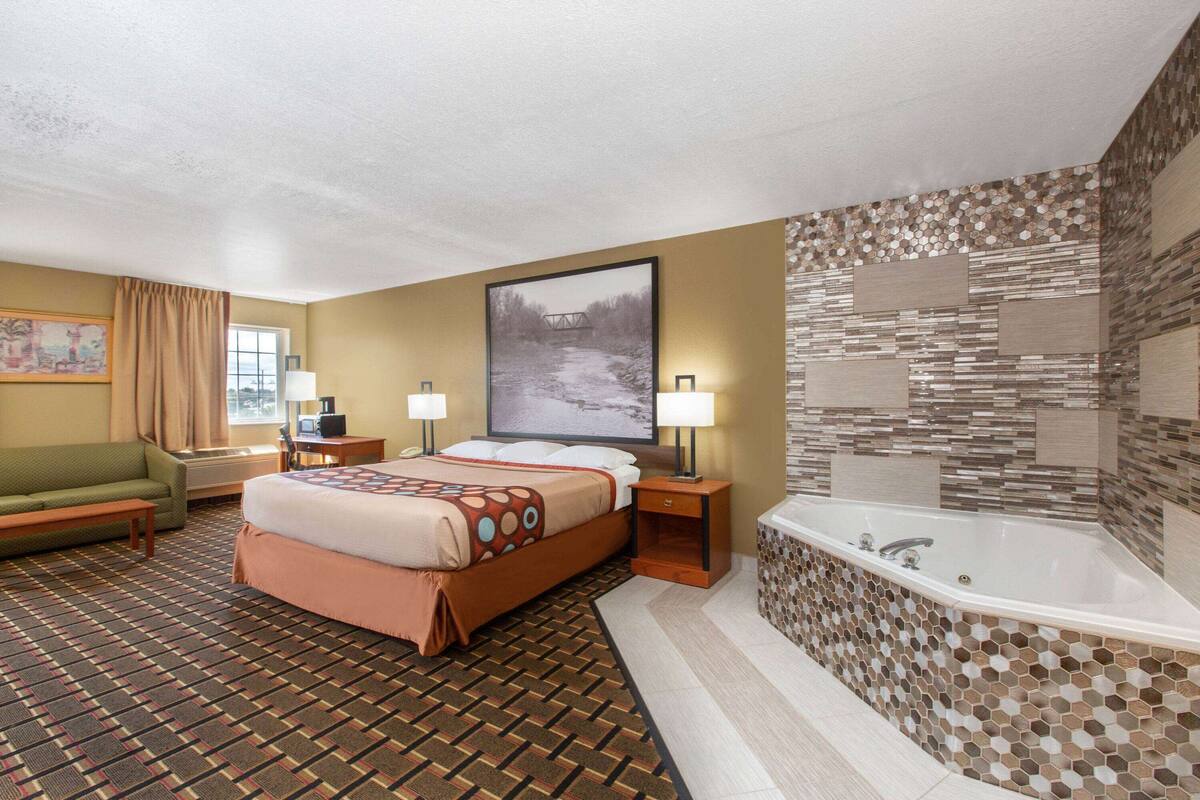 A hotel room with a large bed, a sitting area with a sofa and a table, a desk, a large window, and a bathroom with a bathtub and mosaic tiles.