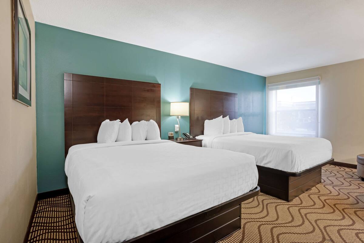 A hotel room with two beds, a nightstand with a lamp, a window with blinds, and a patterned carpet.