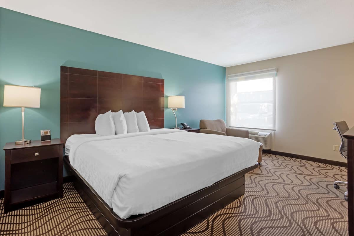 A hotel room with a large bed, two bedside tables, a desk, a chair, and a window with blinds.