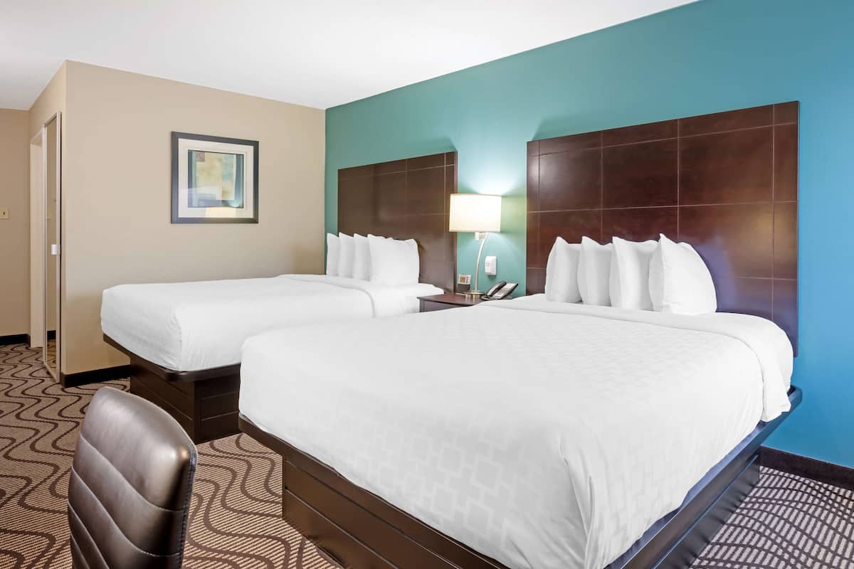A hotel room with two beds, a headboard, a bedside table with a lamp, a chair, and a patterned carpet.