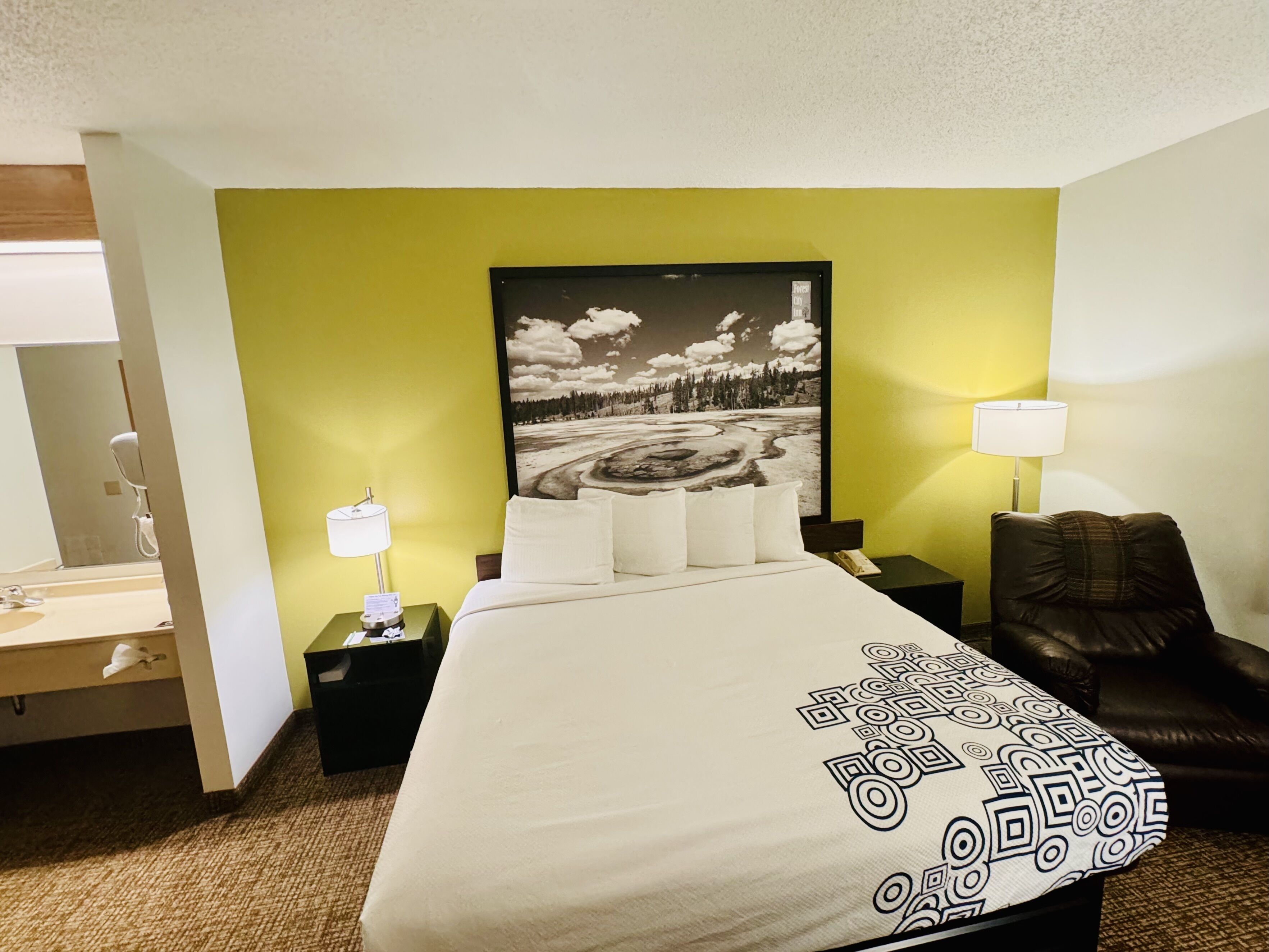 A hotel room with a bed, a nightstand, a lamp, a chair, and a painting on the wall.