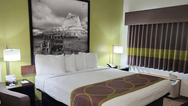 A hotel room with a large bed, two bedside tables with lamps, a chair, a large framed picture on the wall, and a window with curtains. - Forest City Inn (Forest City)