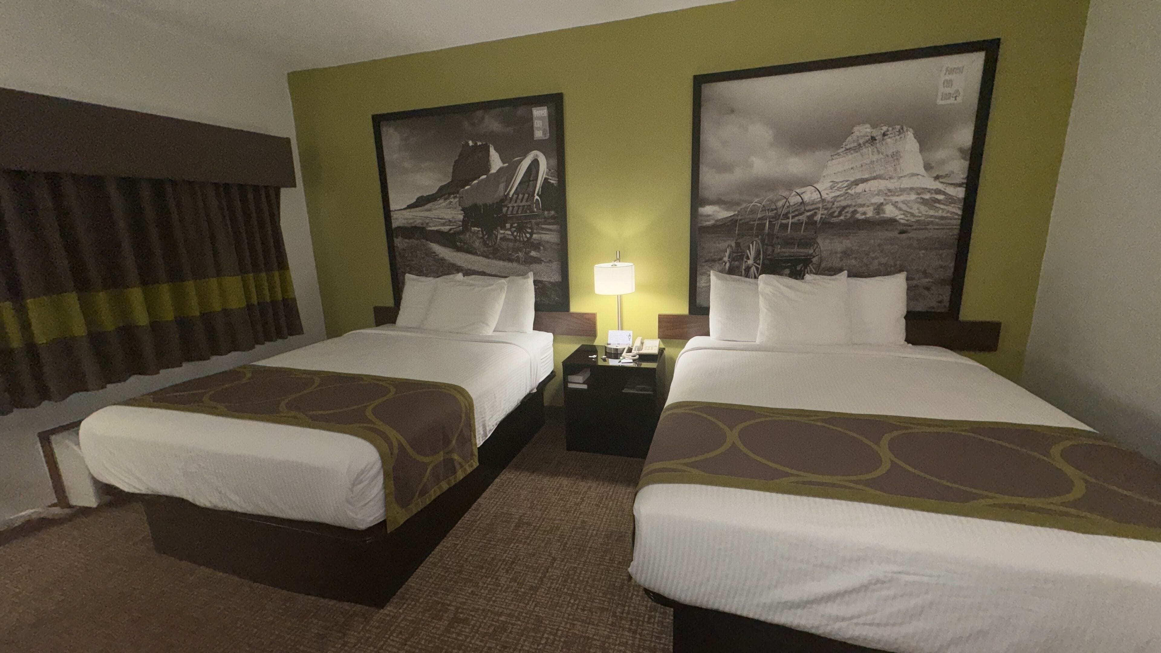 Een hotelkamer met twee bedden, een nachtkastje met een lamp, en twee grote landschapsposters aan de muur.