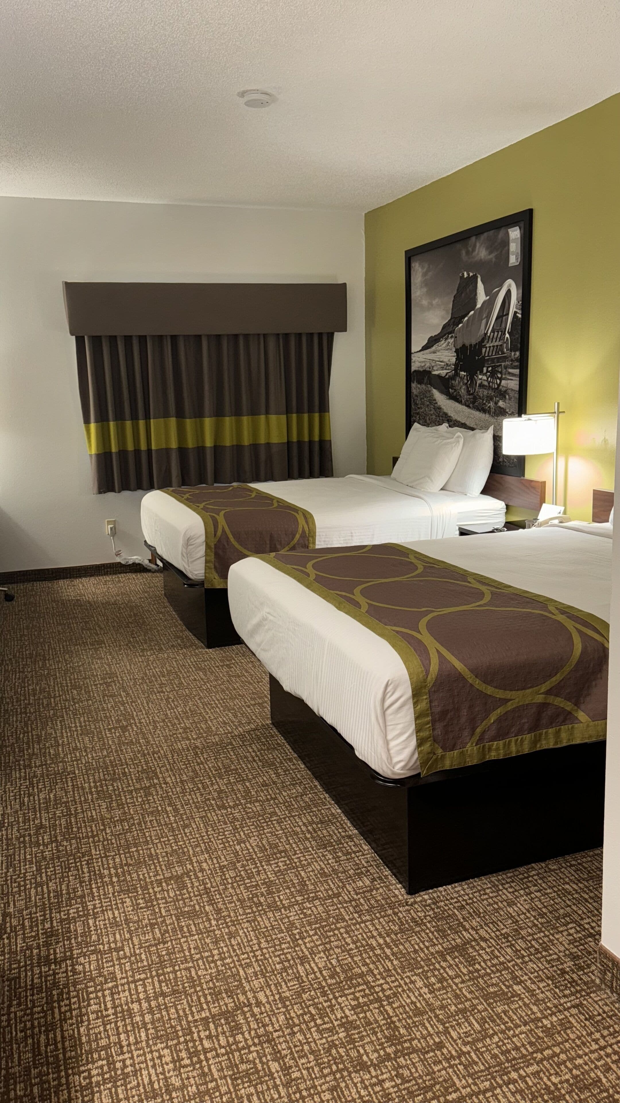 Habitación de hotel con dos camas, un ventanal con cortinas, un cuadro en la pared y suelo alfombrado.