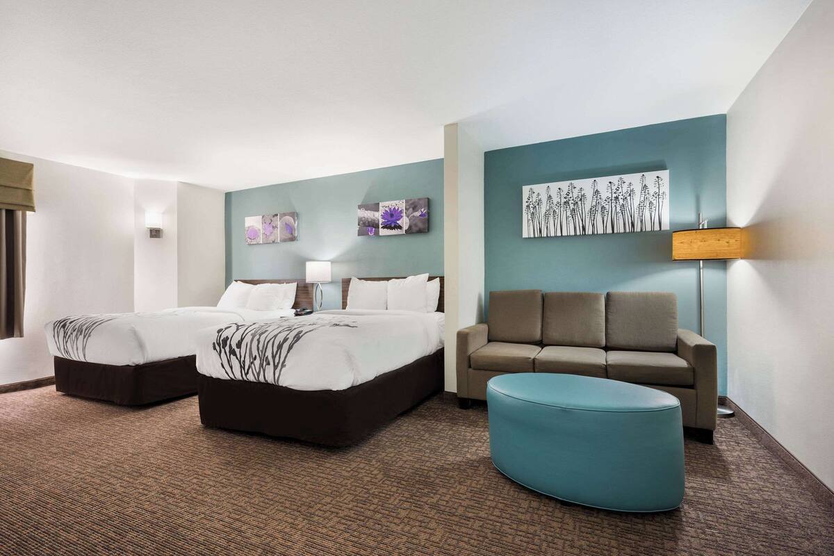 A hotel room with two beds, a sofa, a round ottoman, and artwork on the walls.