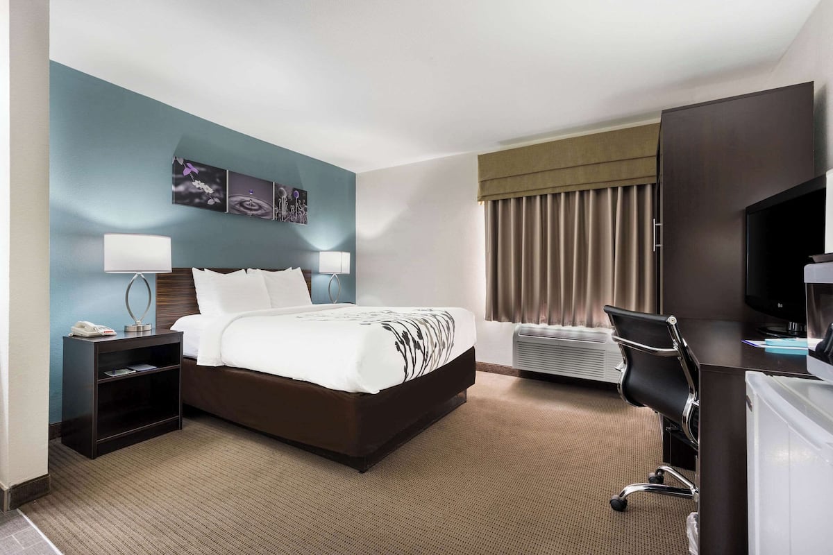 A hotel room with a bed, bedside tables, a desk, a chair, a TV, and a window with curtains.