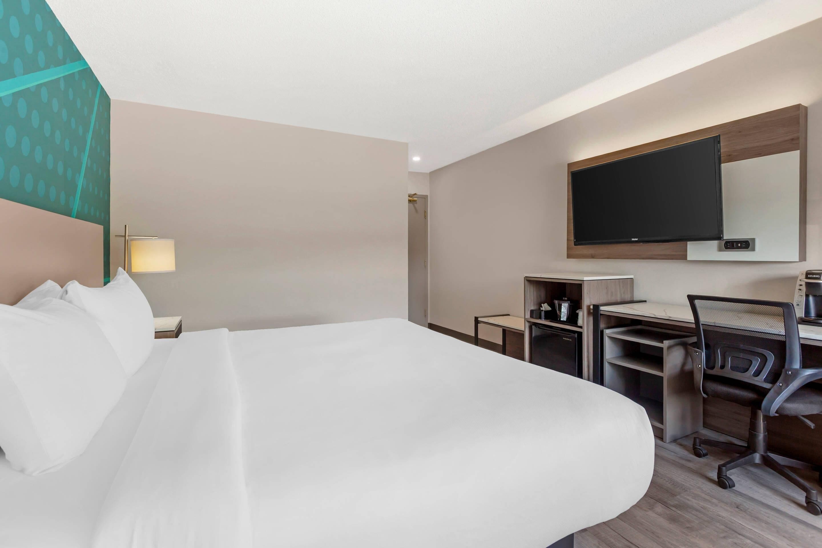 Ein Hotelzimmer mit einem großen Bett, einem Schreibtisch mit Stuhl, einem flachbildfernseher an der Wand und einer Lampe auf dem Nachttisch.