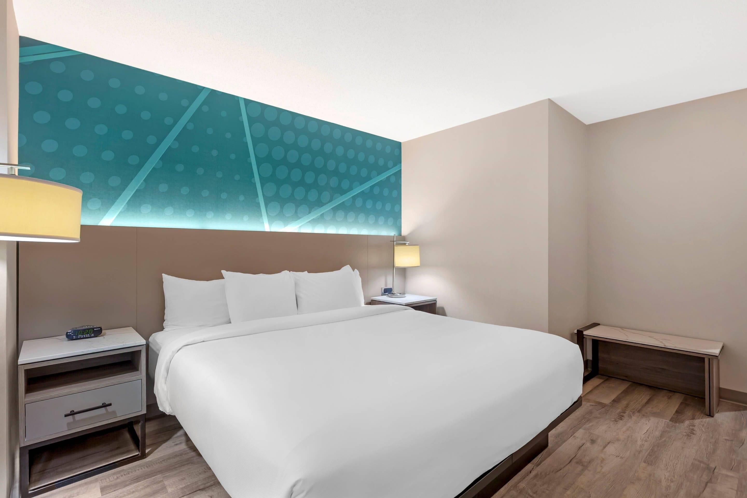 Habitación de hotel con una cama grande, una mesita de noche con lámpara y una pared con diseño geométrico.