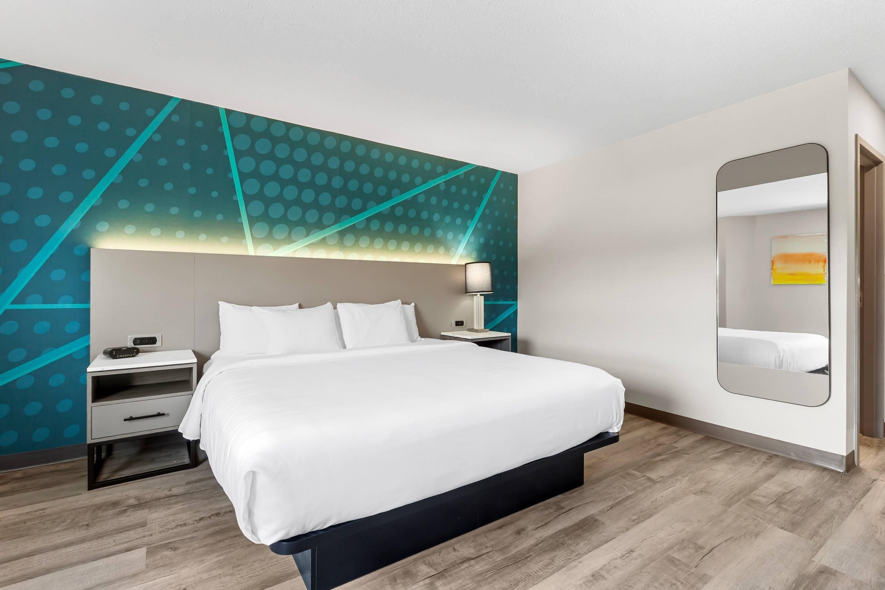 Una habitación de hotel moderna con una cama grande, una mesita de noche, una lámpara y un espejo.
