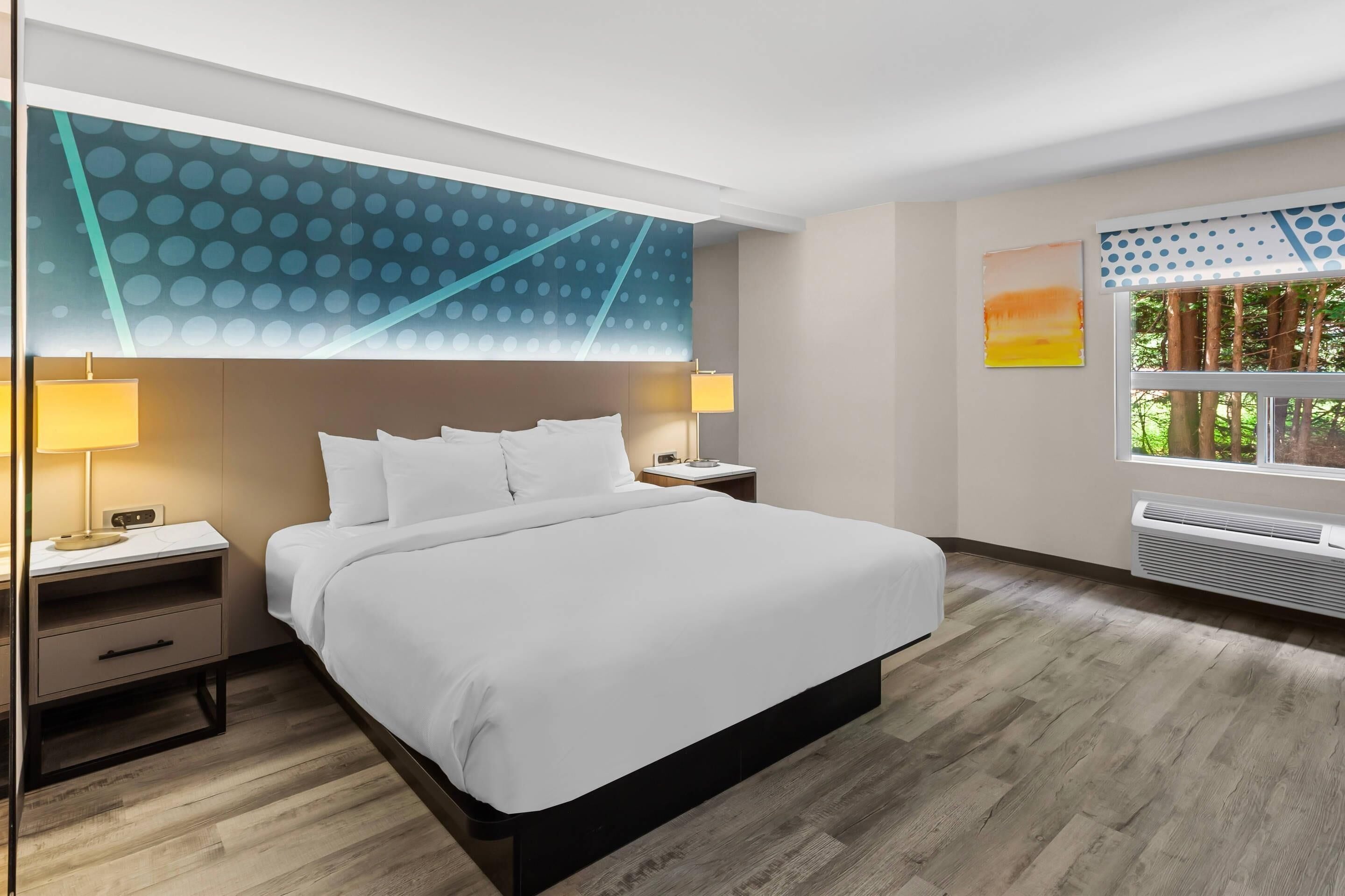 A modern hotel room with a large bed, bedside tables with lamps, a nightstand, a window with a view of trees, and a wall with a geometric pattern.