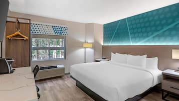 Une chambre d’hôtel avec un grand lit, un bureau, une chaise, un porte-manteau mural, une fenêtre donnant sur des arbres, une lampe fixée au mur et un plafond orné d’un motif géométrique.
