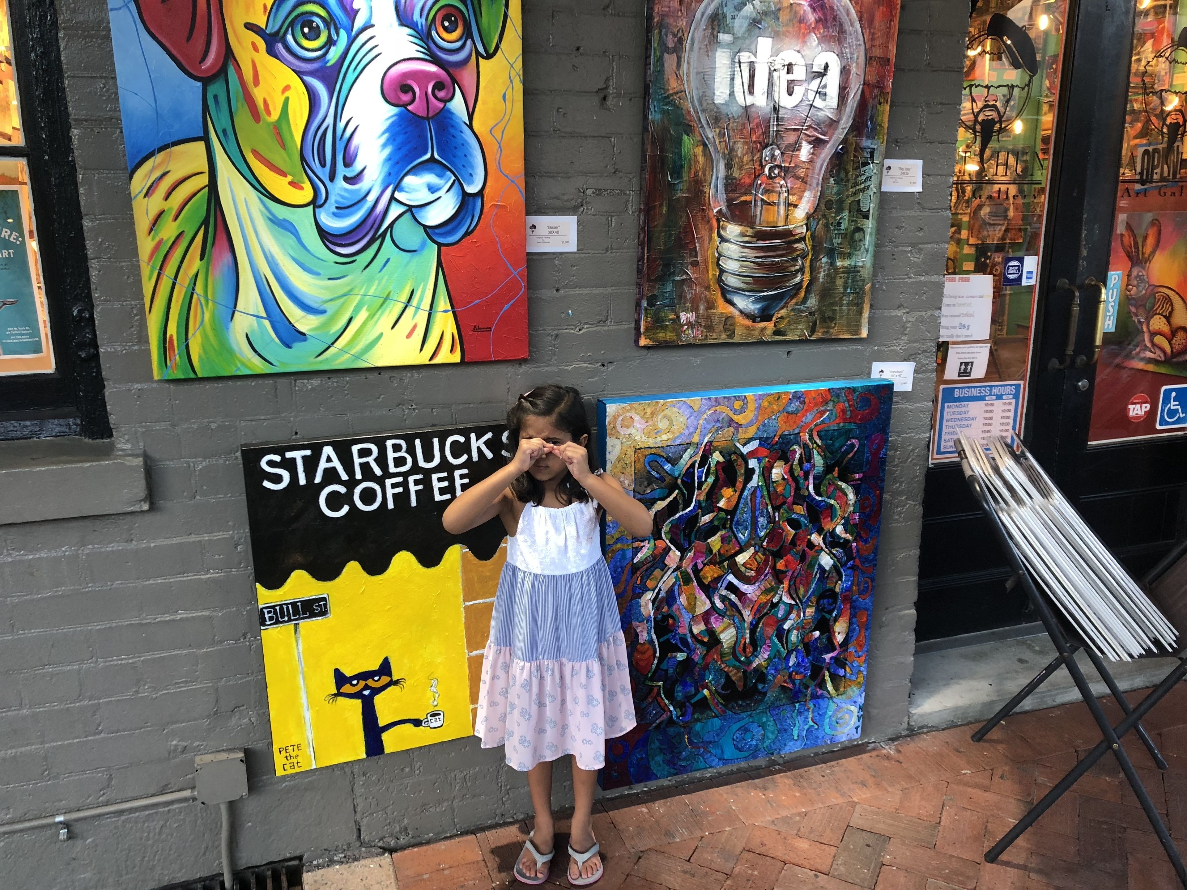 A child standing in front of a wall with various artworks, including a colorful dog, a light bulb with the word 'idea', and a cat illustration.