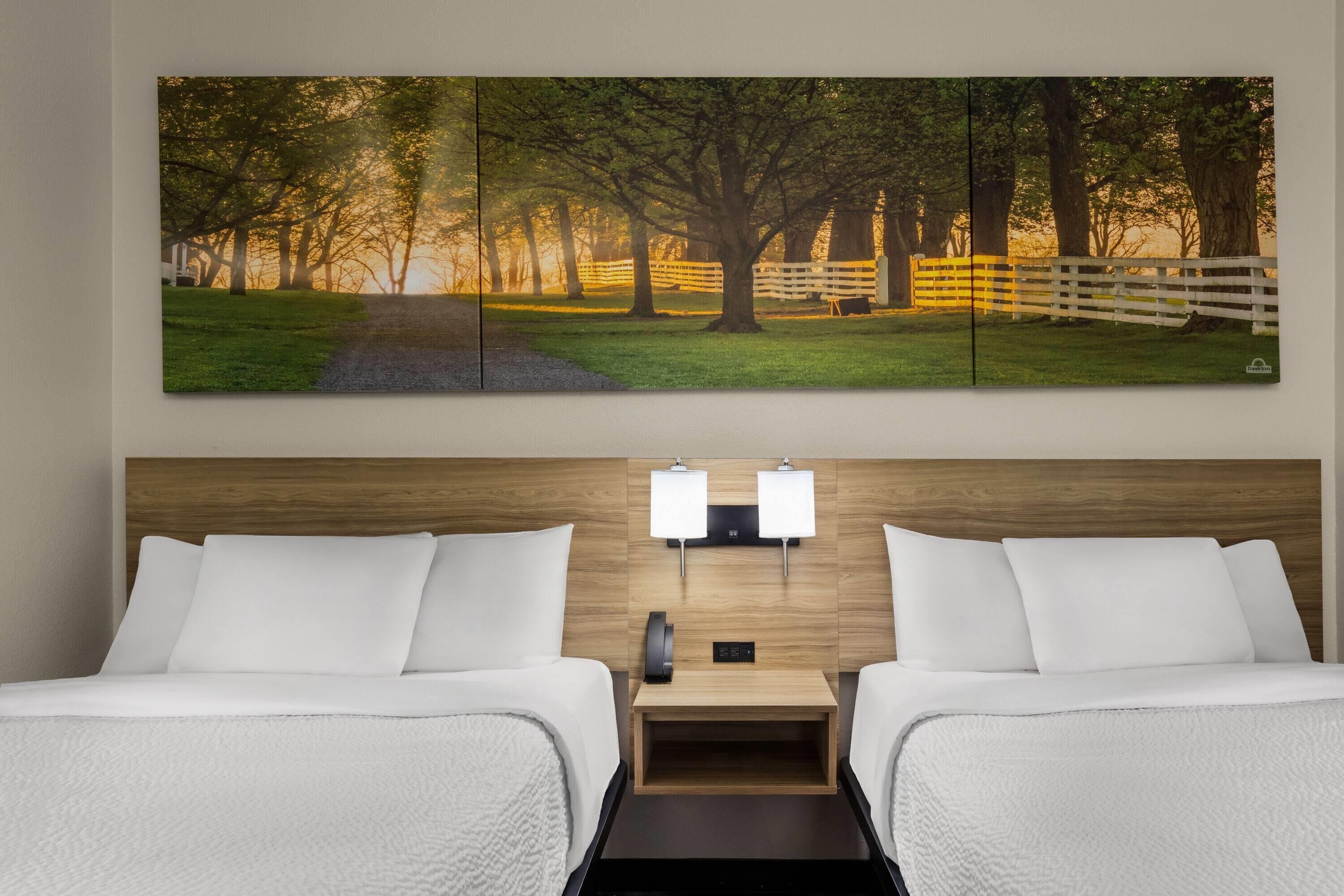 A hotel room with two beds, a headboard with a wooden finish, a nightstand with a lamp, and a large landscape painting above the beds.