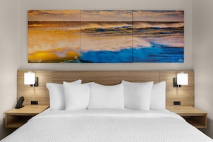 Habitación de hotel con una cama, dos mesitas de noche, una lámpara de pared y un cuadro de un amplio panorama marino.