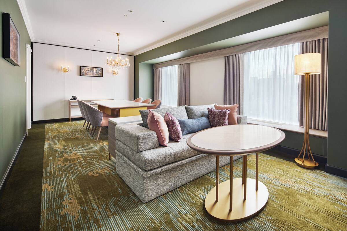 Une chambre d’hôtel moderne avec un canapé gris, une table ronde et un coin repas avec des chaises.