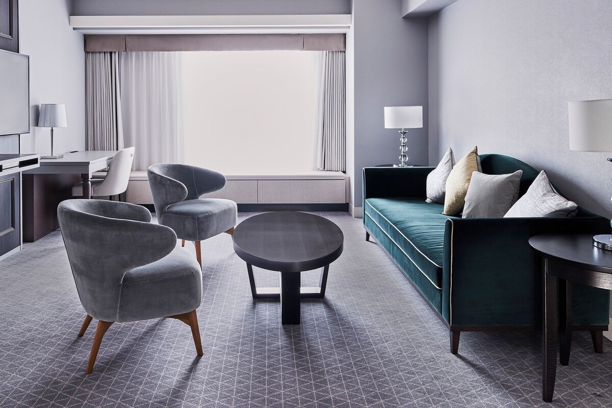 A modern hotel room with a grey sofa, two armchairs, a round coffee table, a TV mounted on the wall, and a large window with curtains.