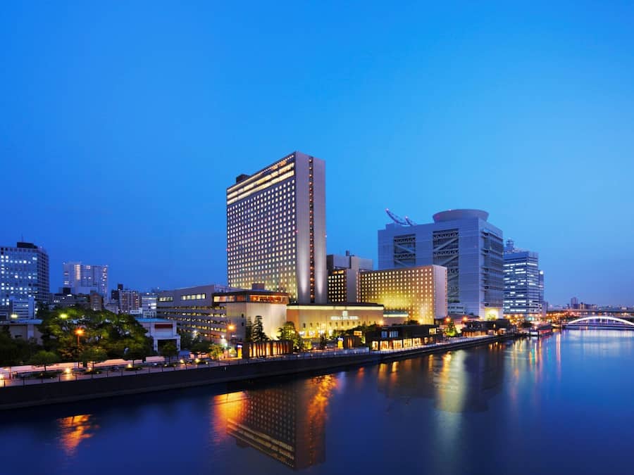 A cityscape at dusk with modern buildings, a river, and a bridge.