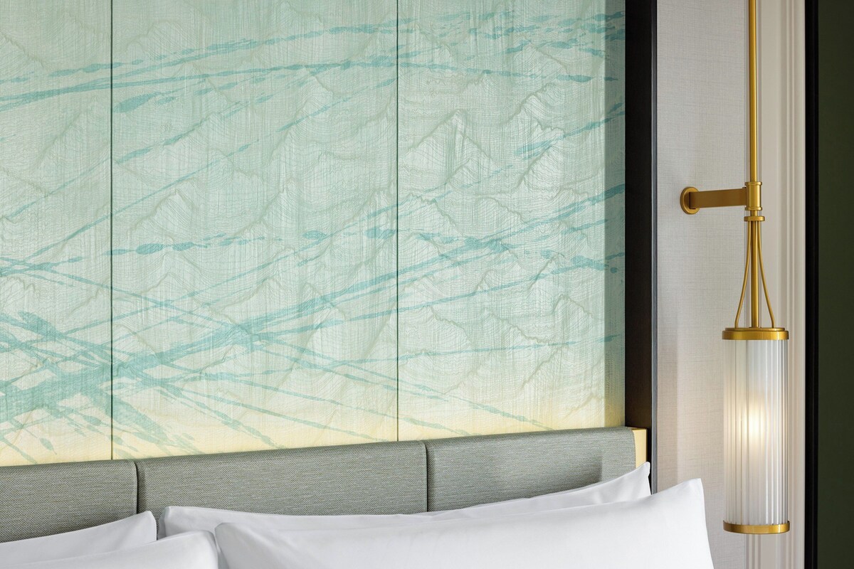 A bed with white linens, a grey headboard, and a wall with a textured abstract design.