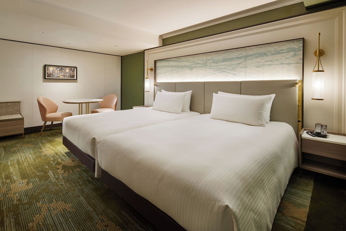 A hotel room with two beds, a small table, a chair, a painting on the wall, and a nightstand with a lamp.