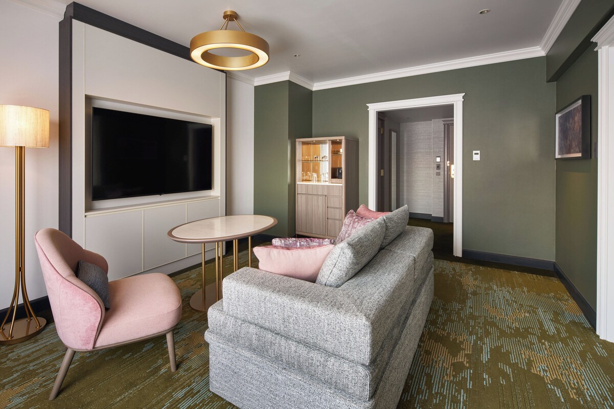 A modern living room with a grey sofa, a pink armchair, a round table, and a large flat-screen TV.
