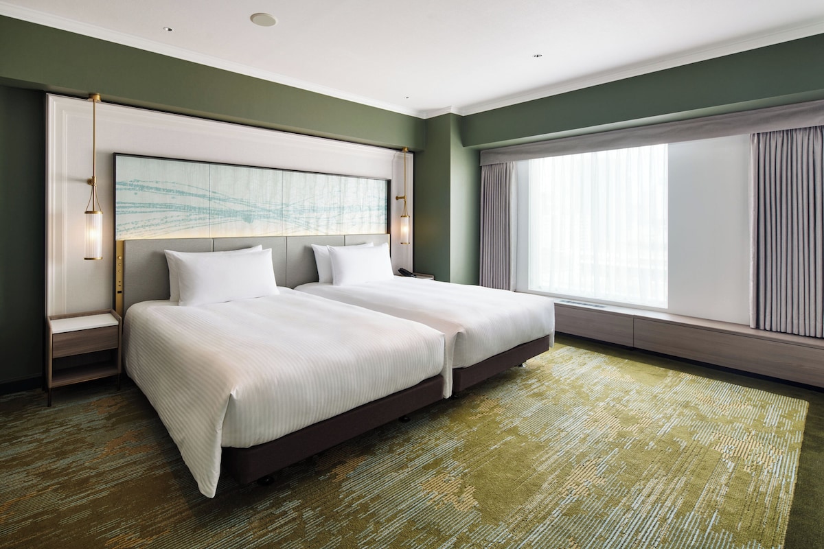 A hotel room with two beds, a large window with curtains, a headboard with artwork, and a carpeted floor.