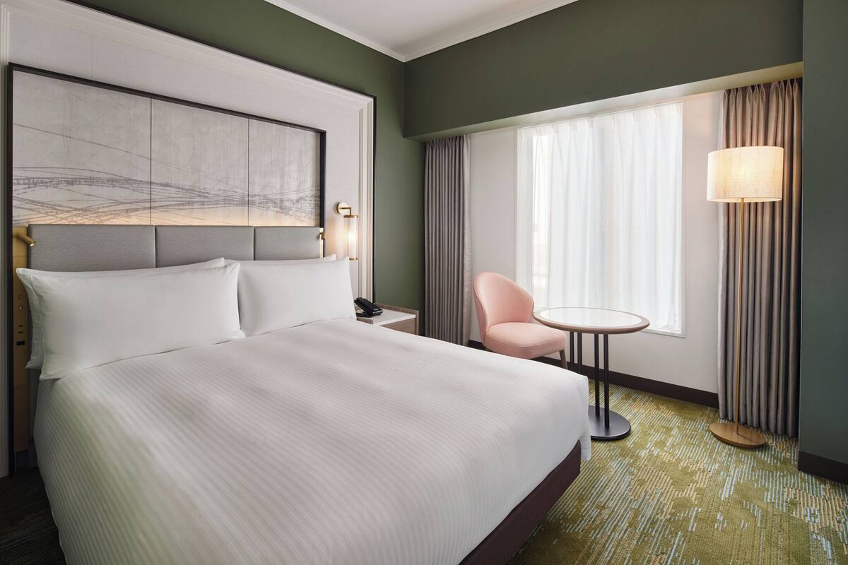 Une chambre d’hôtel avec un grand lit, une petite table ronde, une chaise, un bureau avec une lampe et une grande œuvre d’art accrochée au-dessus de la tête de lit.