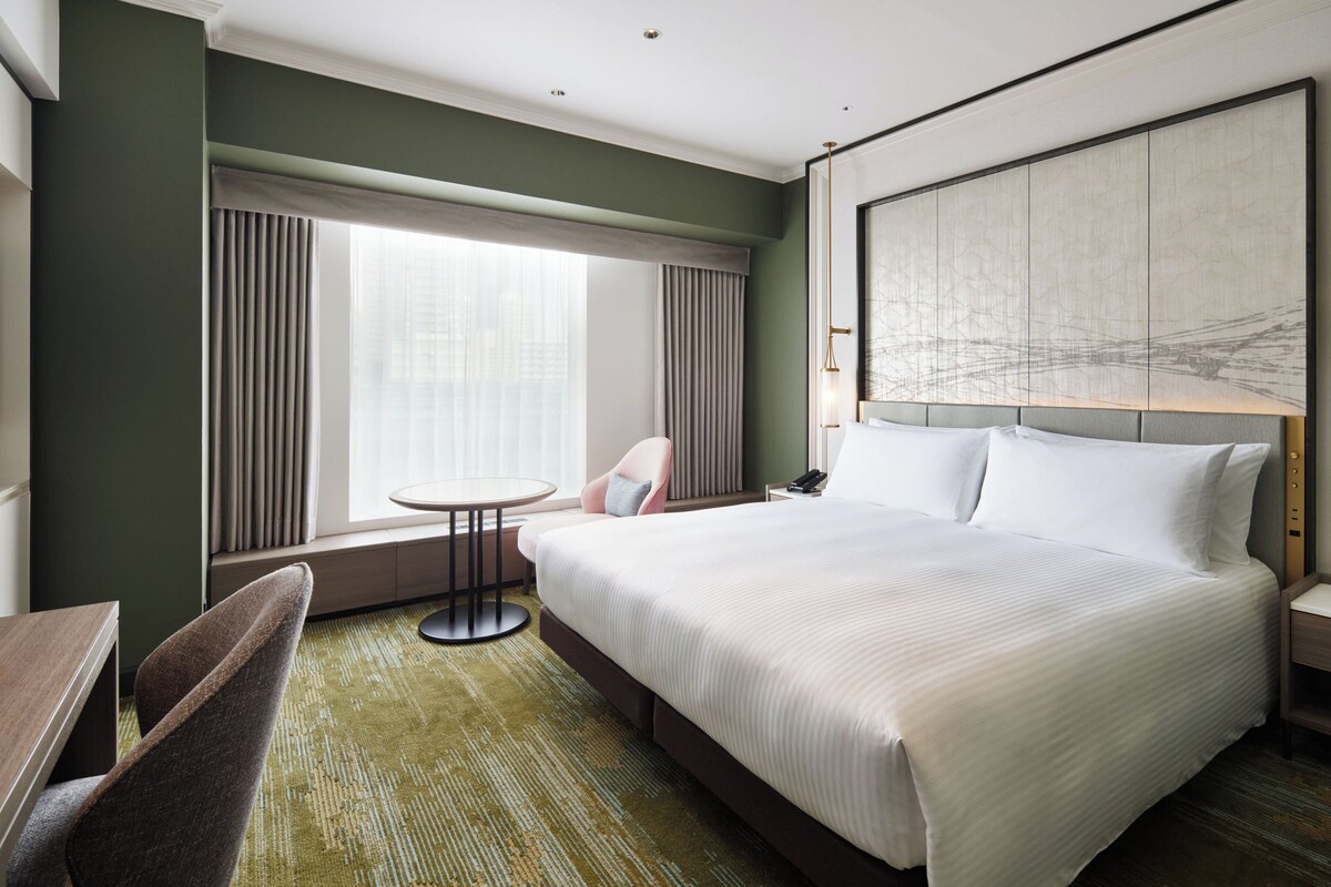A hotel room with a large bed, a small round table, a chair, and a window with curtains.