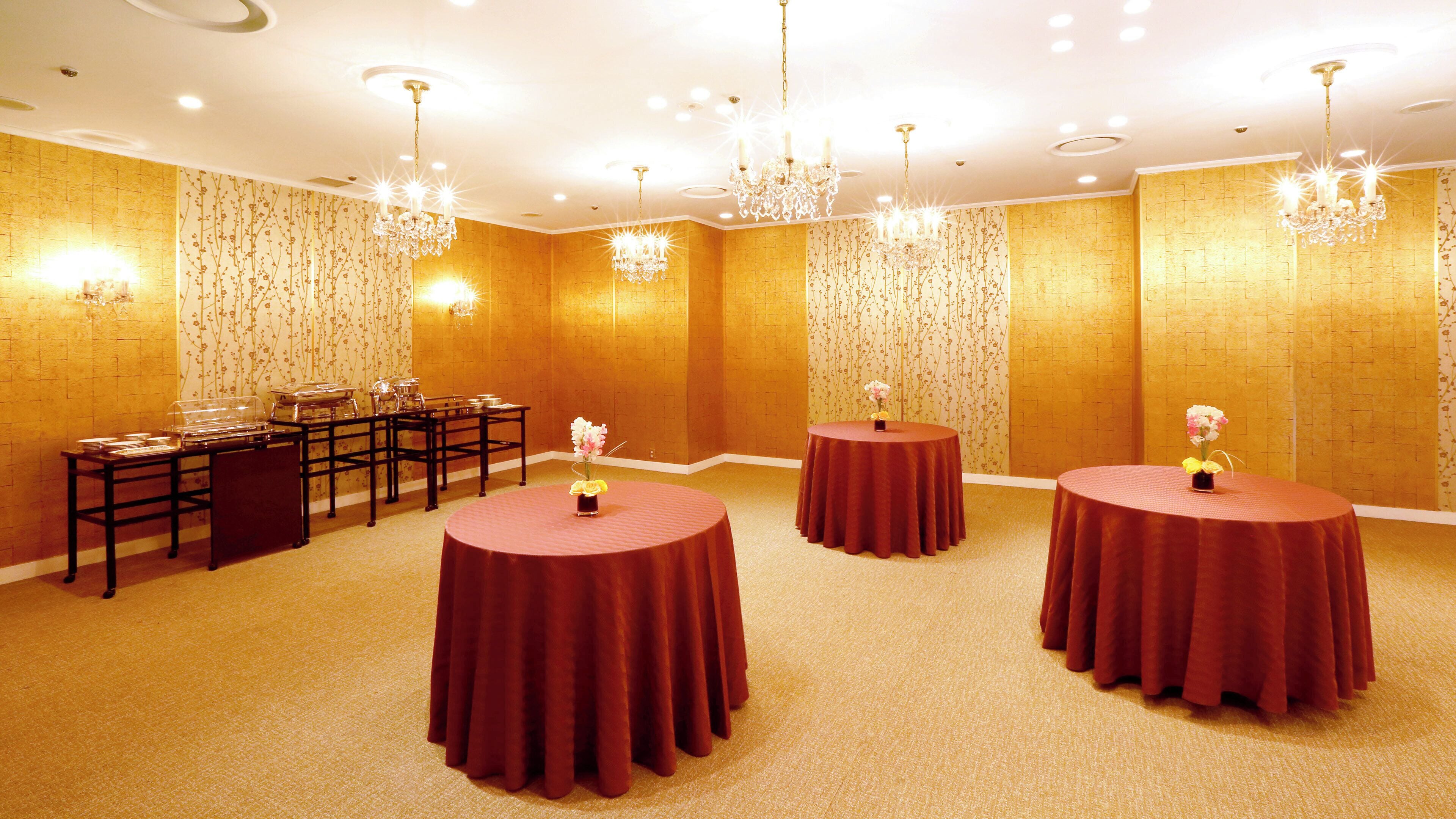 A conference room with four round tables, each with a red tablecloth and a vase of flowers. There are chandeliers hanging from the ceiling, and a buffet table with glassware on the left.