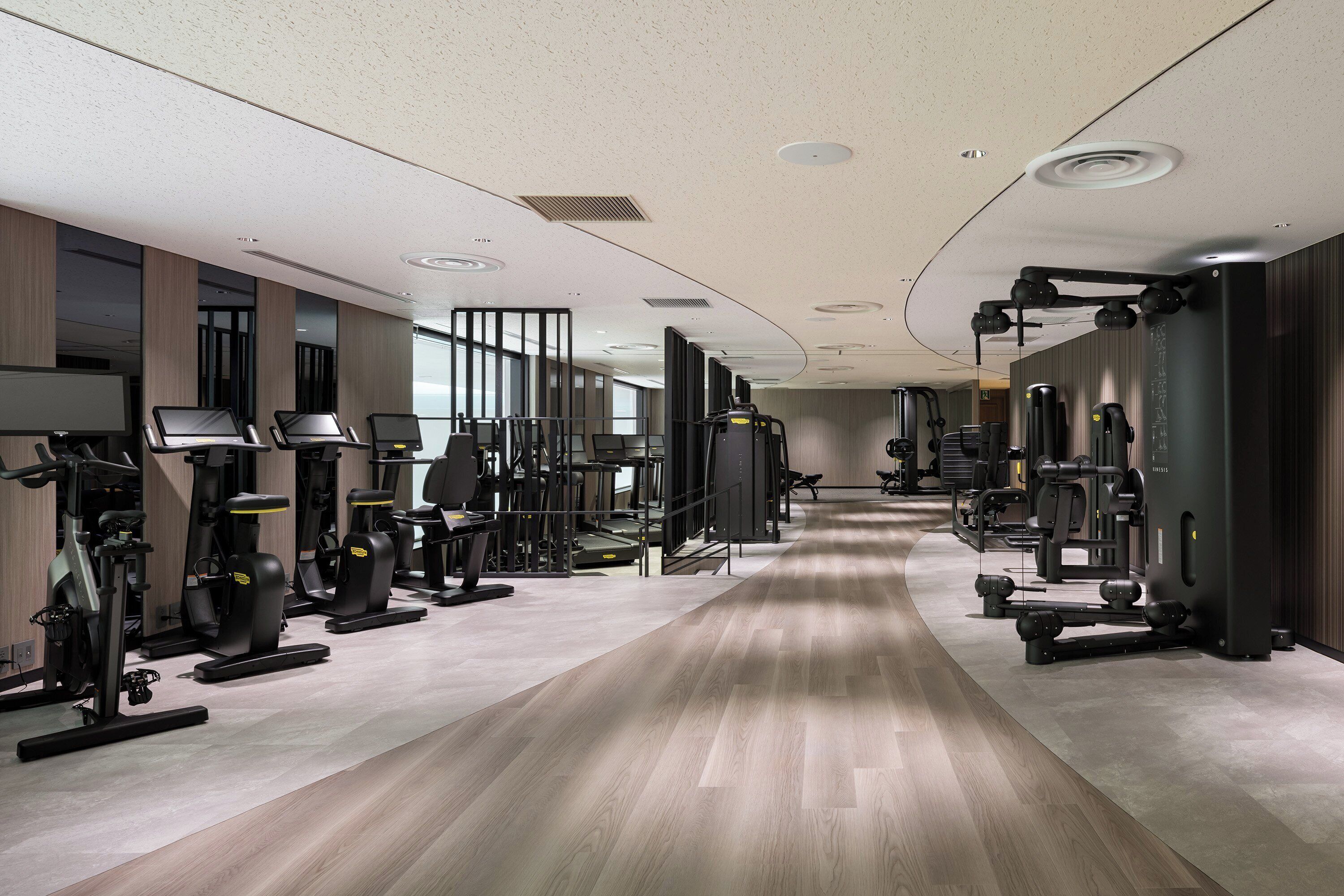 A modern gym with various exercise machines, a long wooden floor, and a spacious layout.